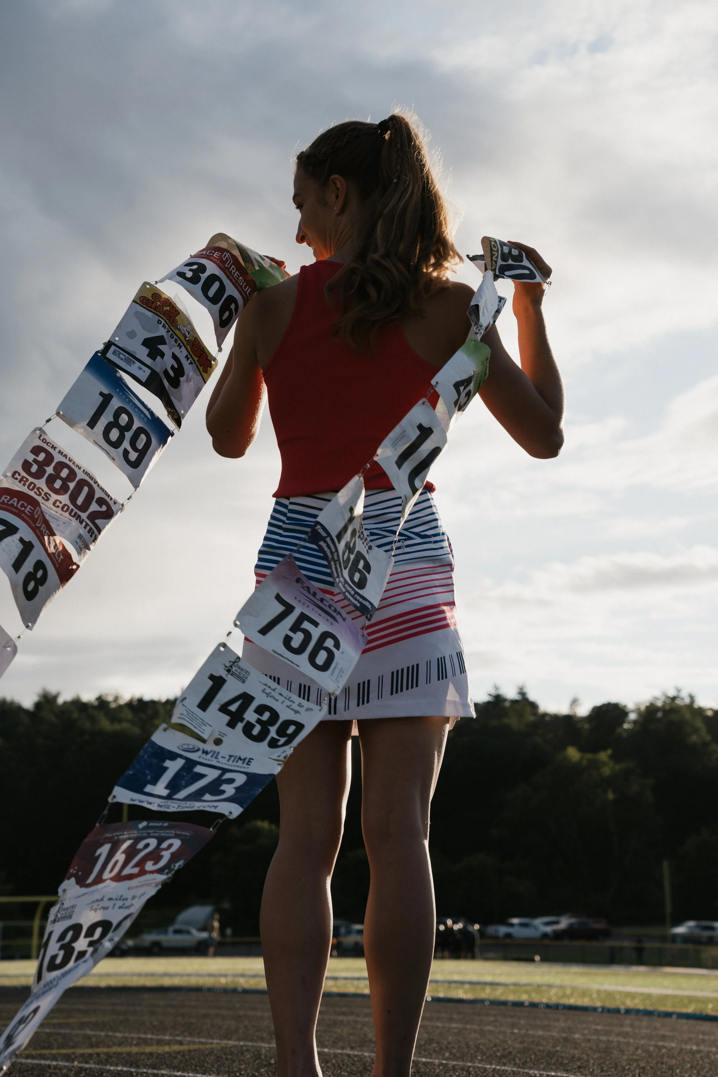 A woman with a ponytail in athletic clothing is placing race bibs on her body at a track.