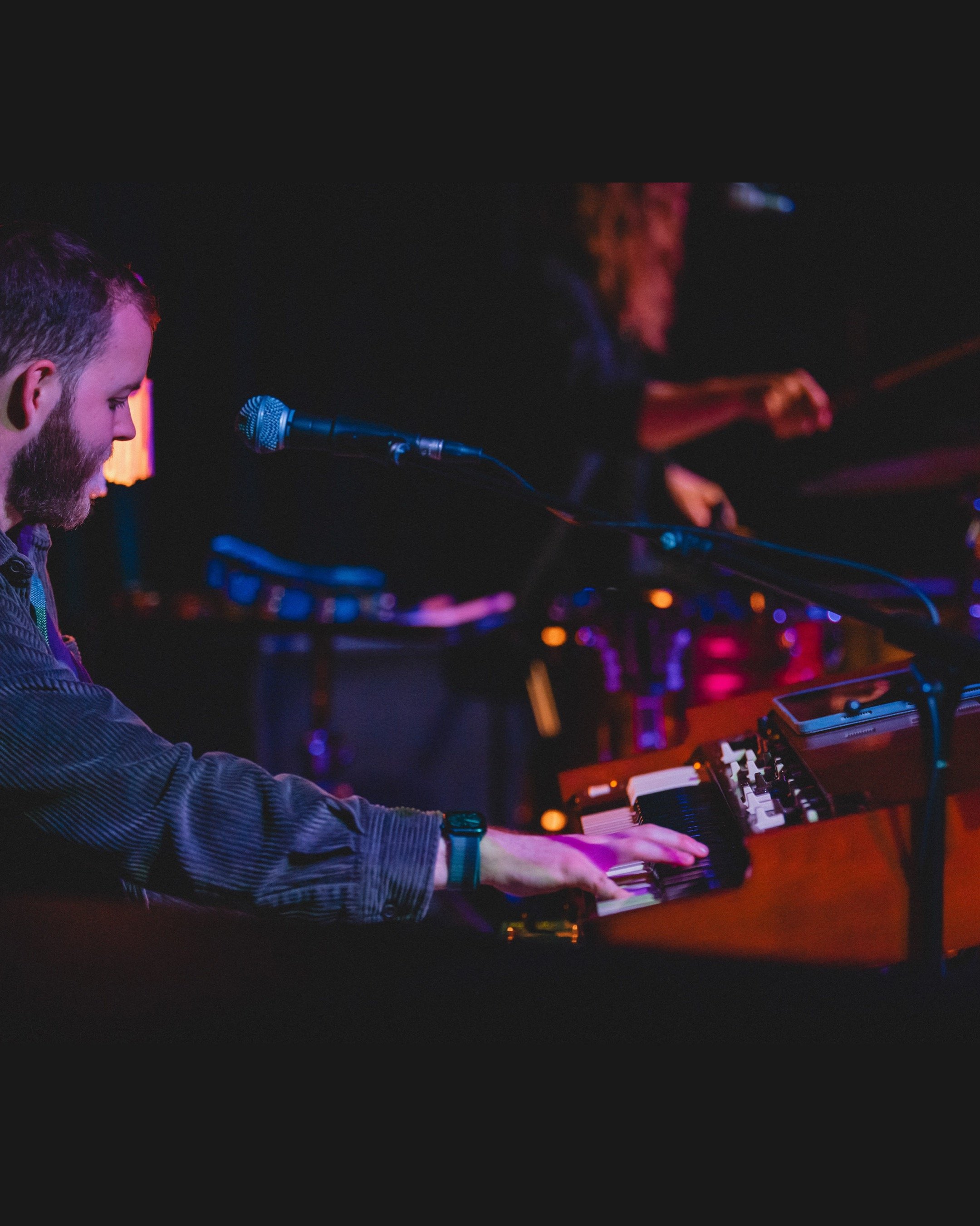 A musician playing a keyboard on stage illuminated with purple and pink lights.