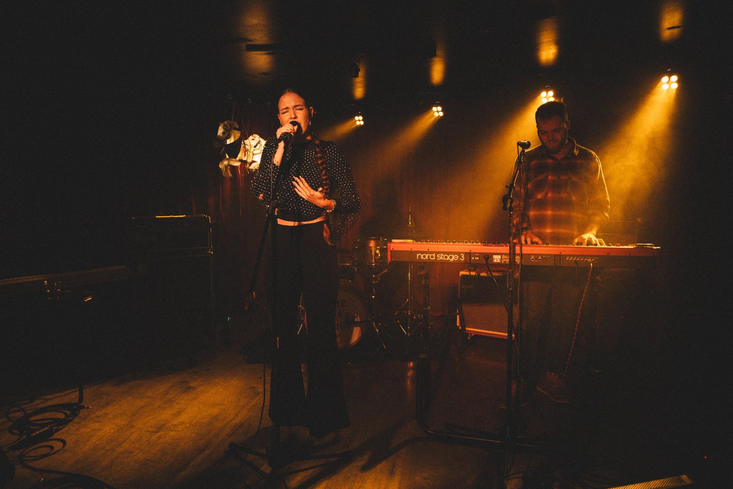 A woman singing with a microphone on stage in front of a keyboardist, during a live music performance with warm, yellow lighting.