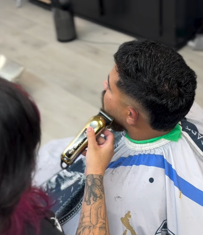 Man getting a gold hair clipper haircut at a barbershop, with a barber holding the clipper and working on his beard.
