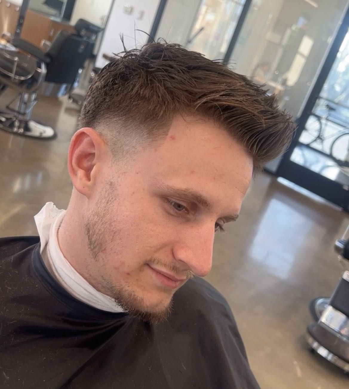 Young man getting a haircut in a salon with modern décor and large windows.