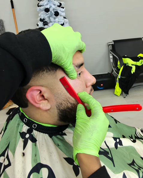 A barber is cutting a man's hair with a straight razor, wearing green gloves.