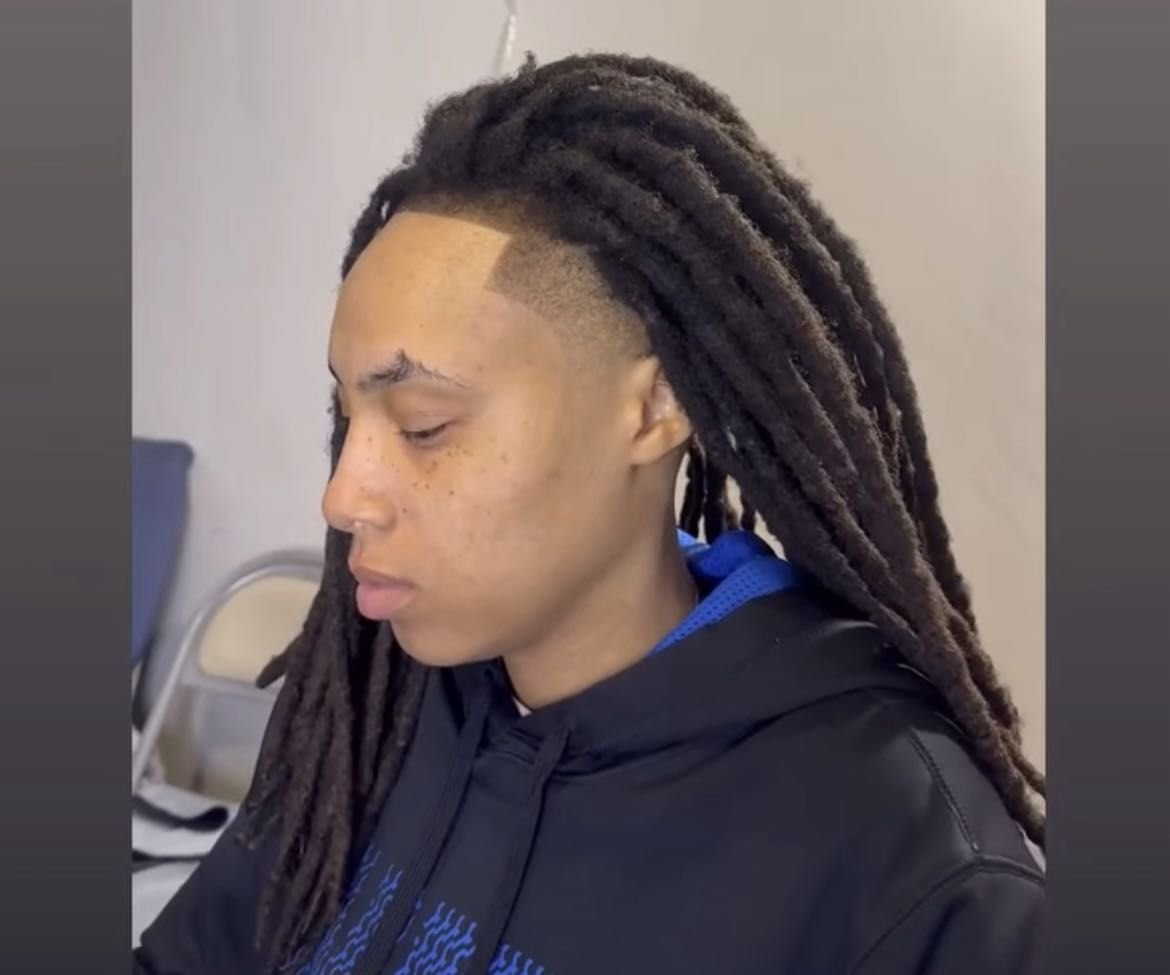 A person with long dreadlocks, wearing a black hoodie, looking downward.