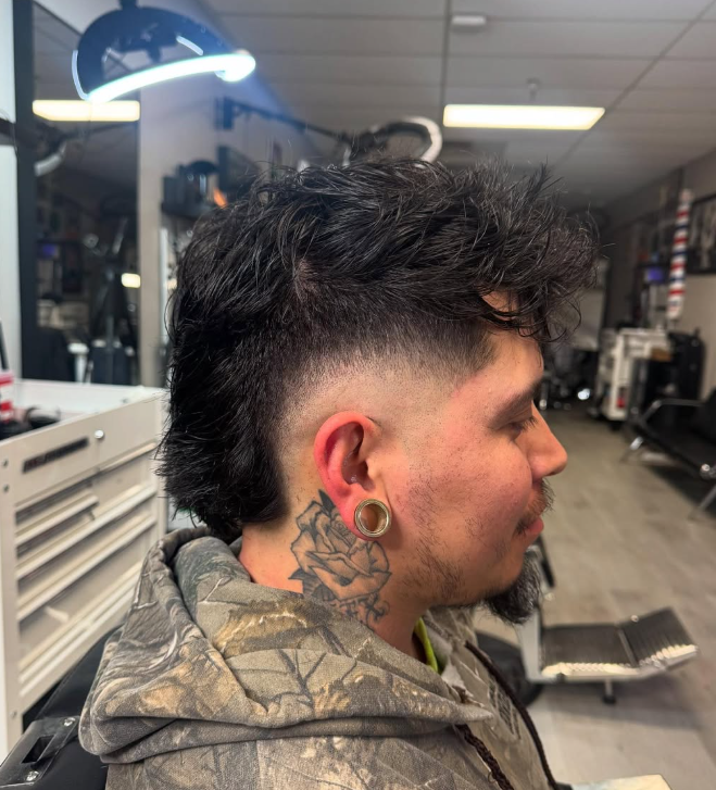 A man in a salon with a mohawk haircut, featuring a fade on the sides and longer curly hair on top, sitting in a barber chair.