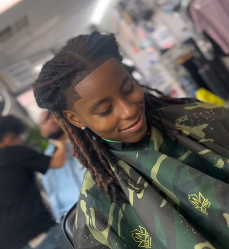 Woman with dreadlocks in a salon chair, wearing a camouflage haircutting cape, smiling with eyes closed.