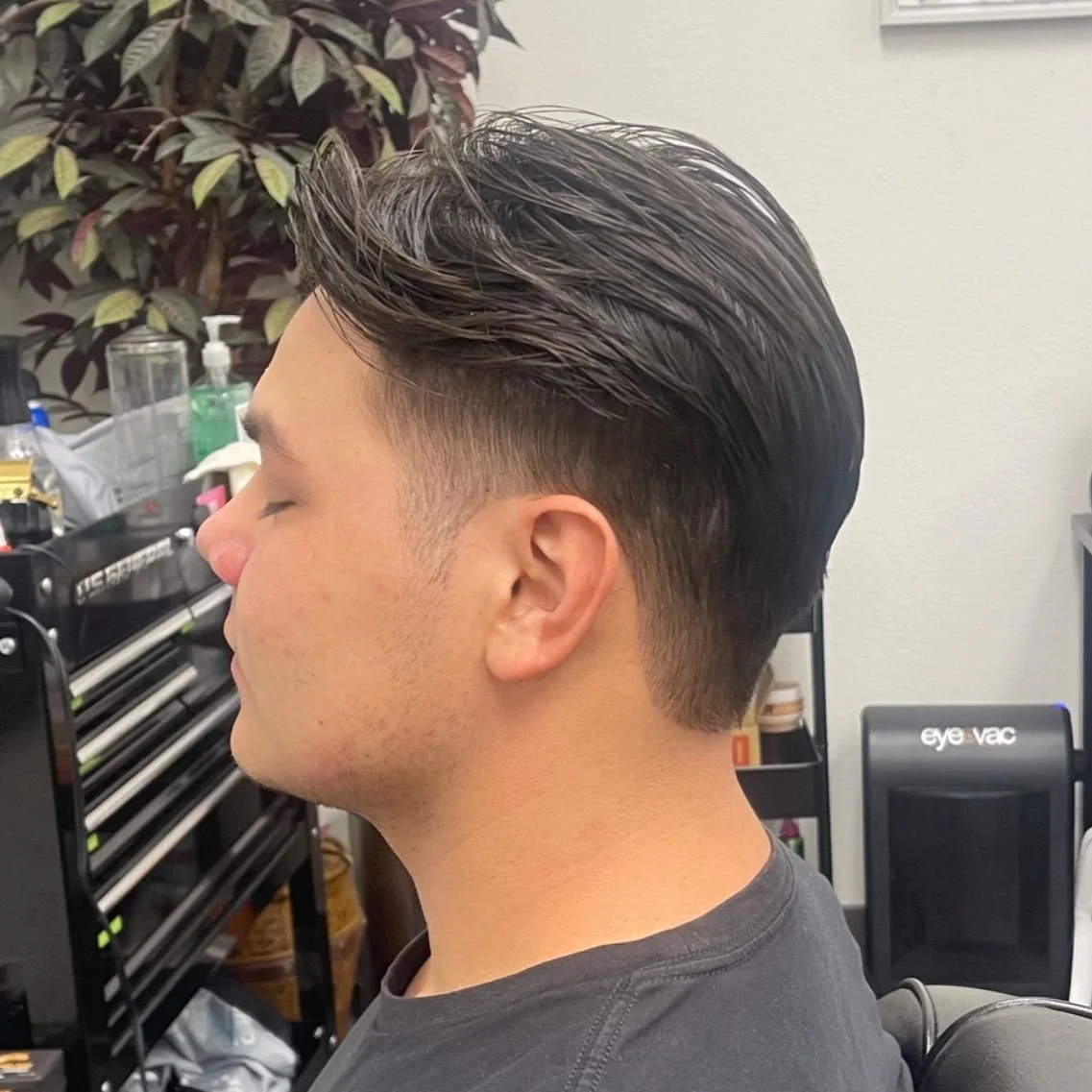 A man with dark, slicked-back hair sitting with his eyes closed in a barber shop, with hair products and tools visible in the background.