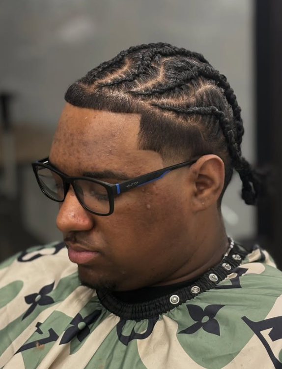A person with braided hair, wearing glasses and a patterned cape, sitting in a barbershop.