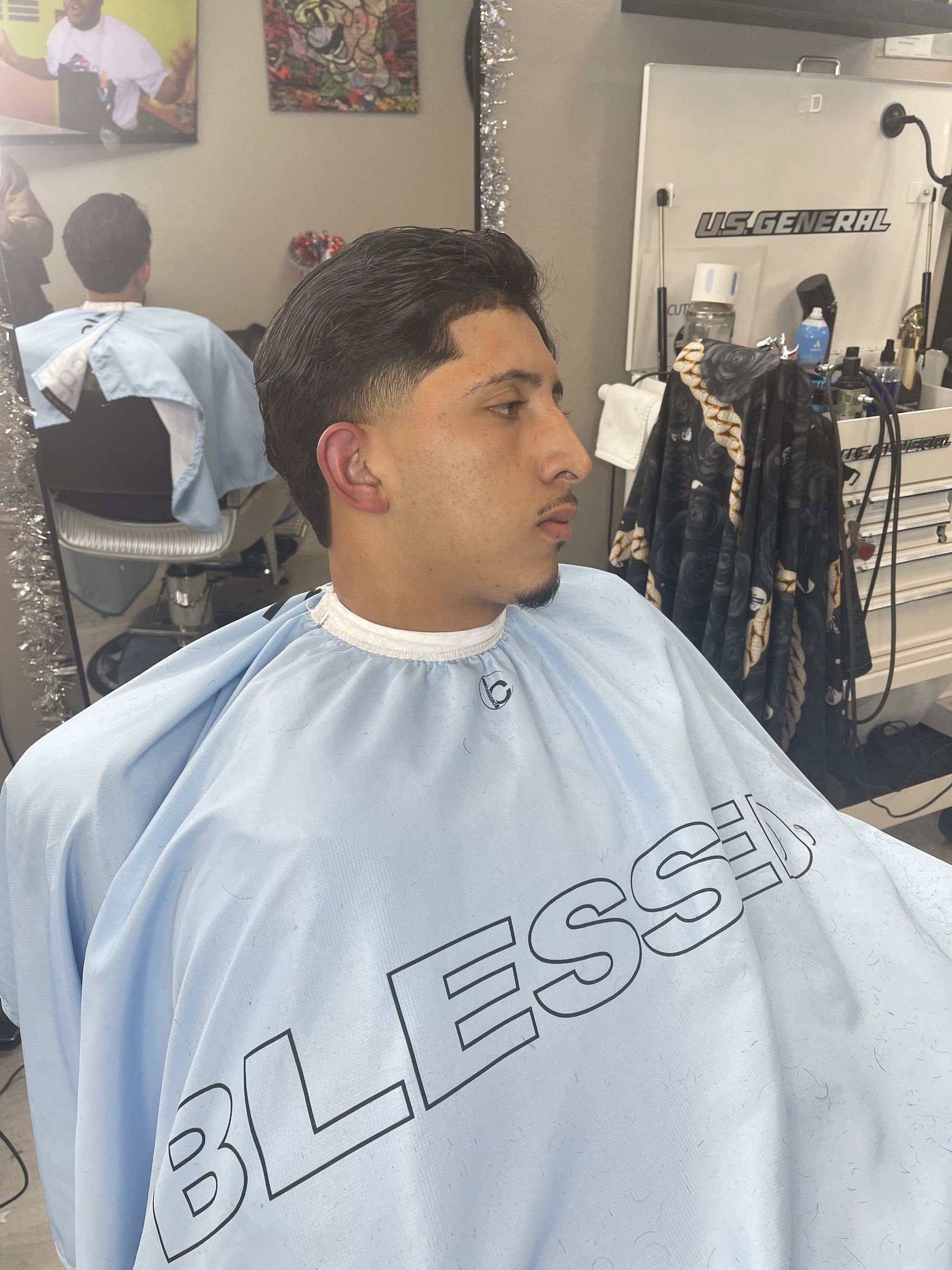 A man getting a haircut in a salon, wearing a light blue barber cape with the word 'BLESSED' on it, and a backdrop of barber equipment and wall decor.