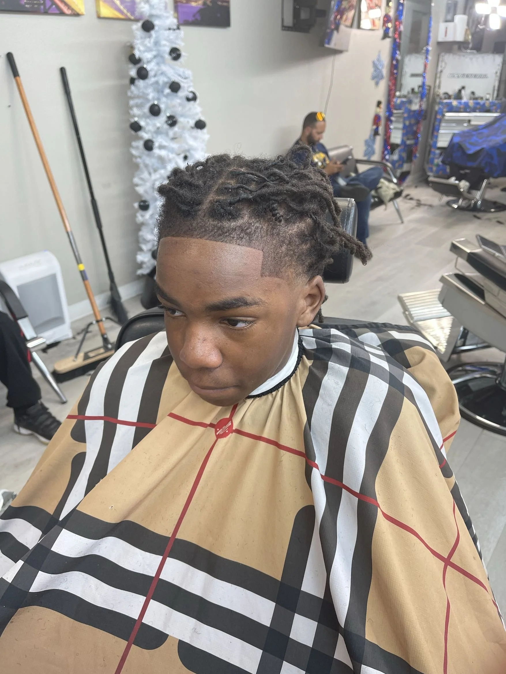 Young man sitting in a barber shop with a freshly styled haircut, wearing a patterned barber cape, and a white Christmas tree in the background.