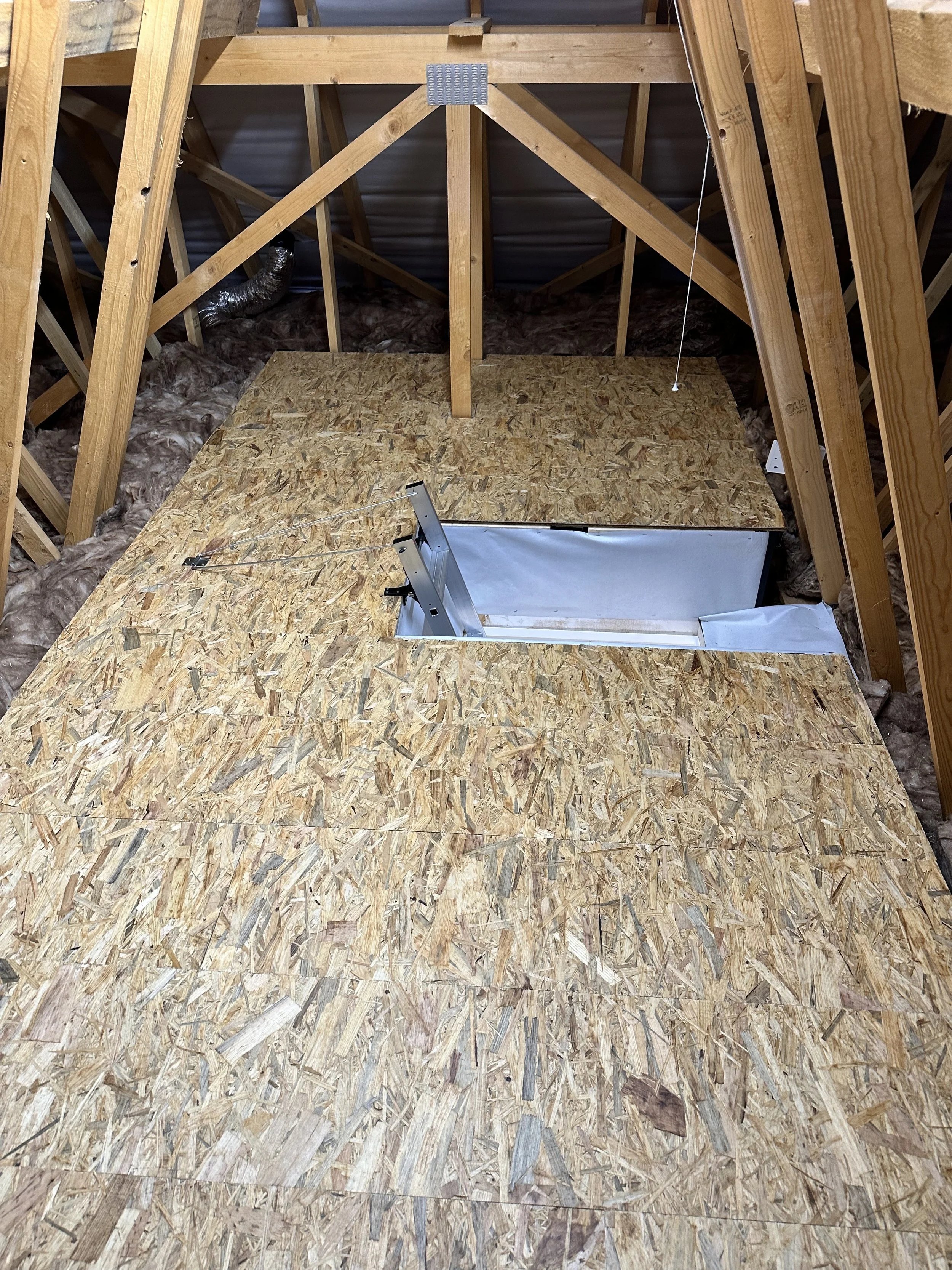 The image shows an attic with exposed wooden framing, insulation, and a partially installed hatch or opening in the raised floor.