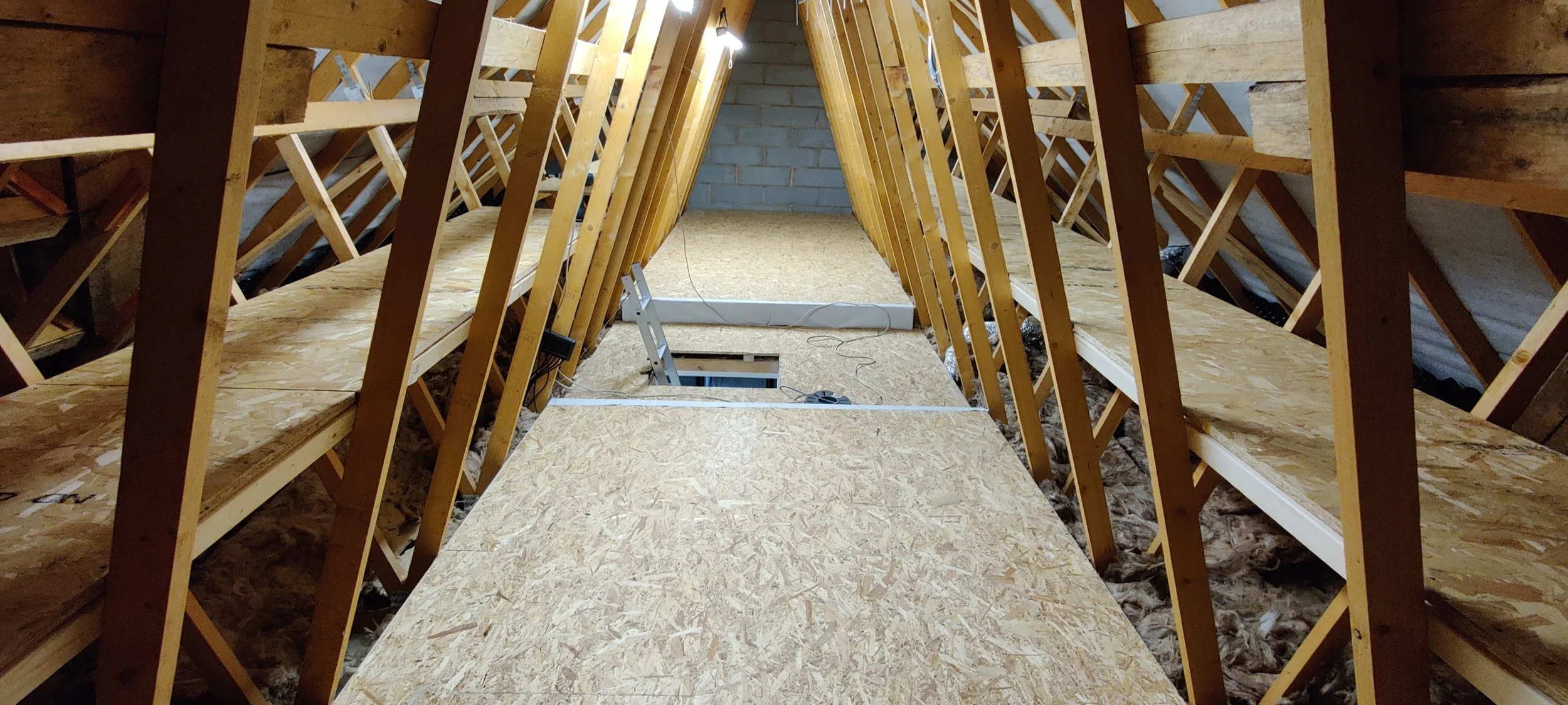 Attic space under construction with exposed wooden framing and OSB flooring, some electrical wiring, and a ladder.