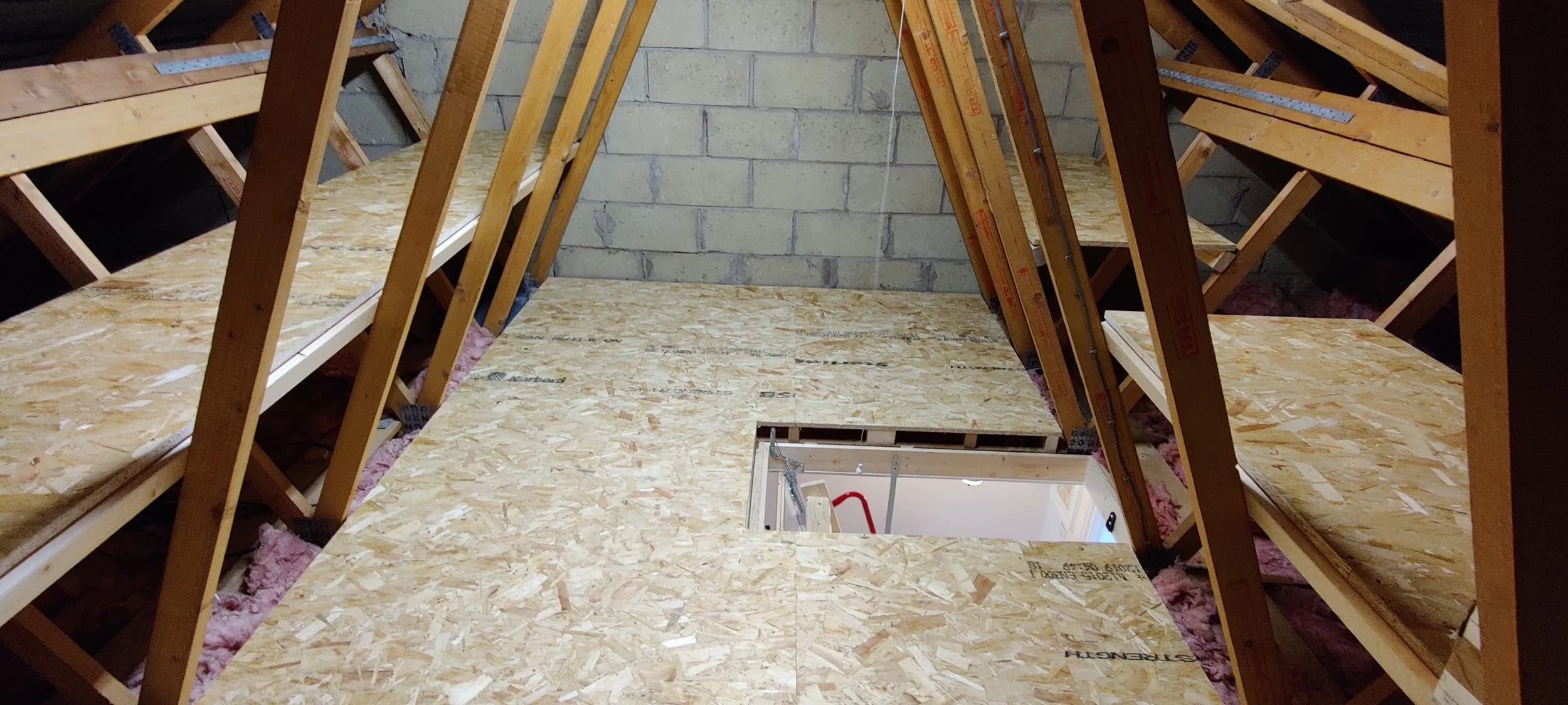 Attic under construction with plywood flooring and exposed wooden framing, staircase opening in the center, brick wall at the back, and pink insulation visible beneath the floorboards.