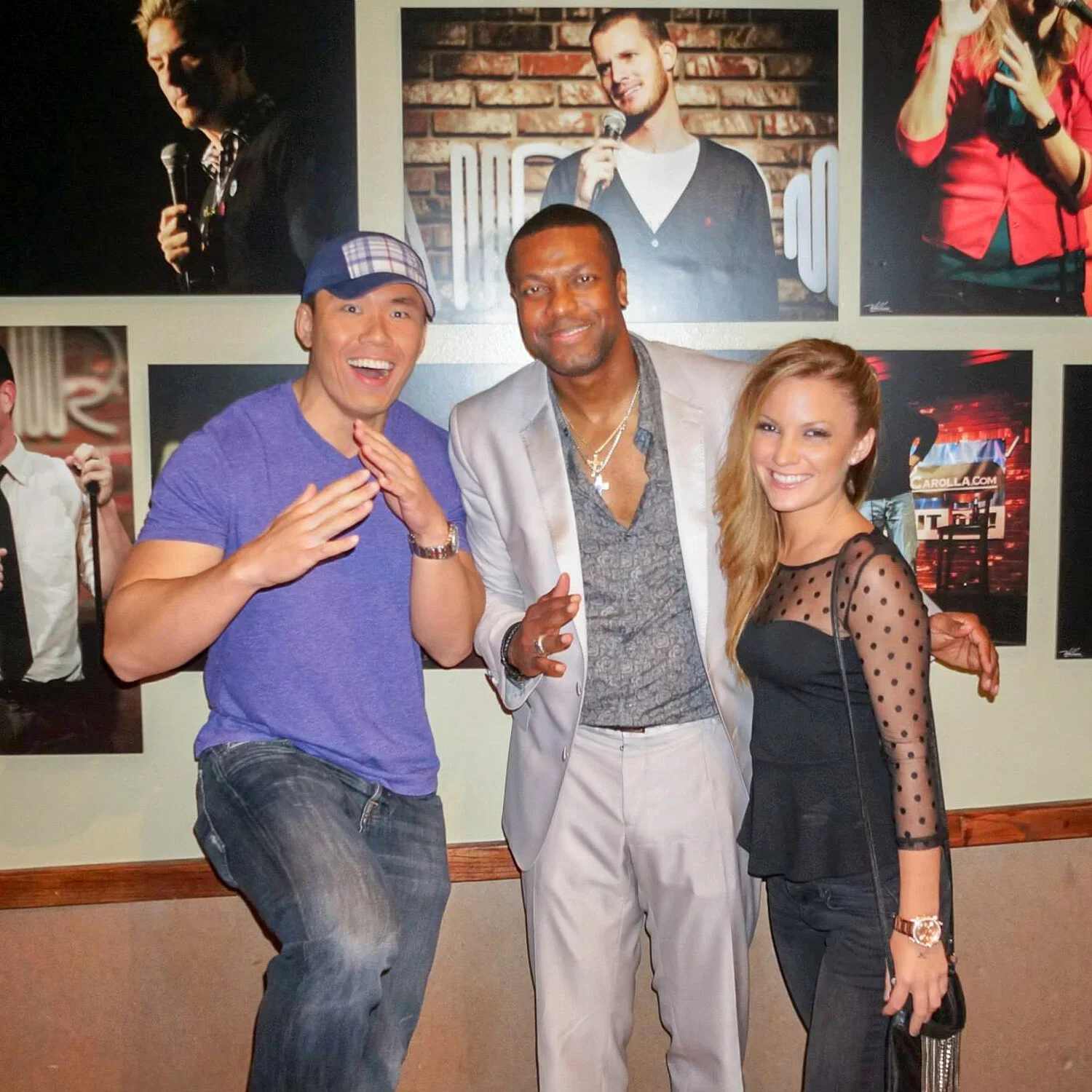 A man in a purple t-shirt and checkered cap smiling and placing his hand near his chin. In the middle is a man in a light gray suit with a patterned shirt and necklaces, smiling and raising his hand. To his right is a woman with long blonde hair, wea