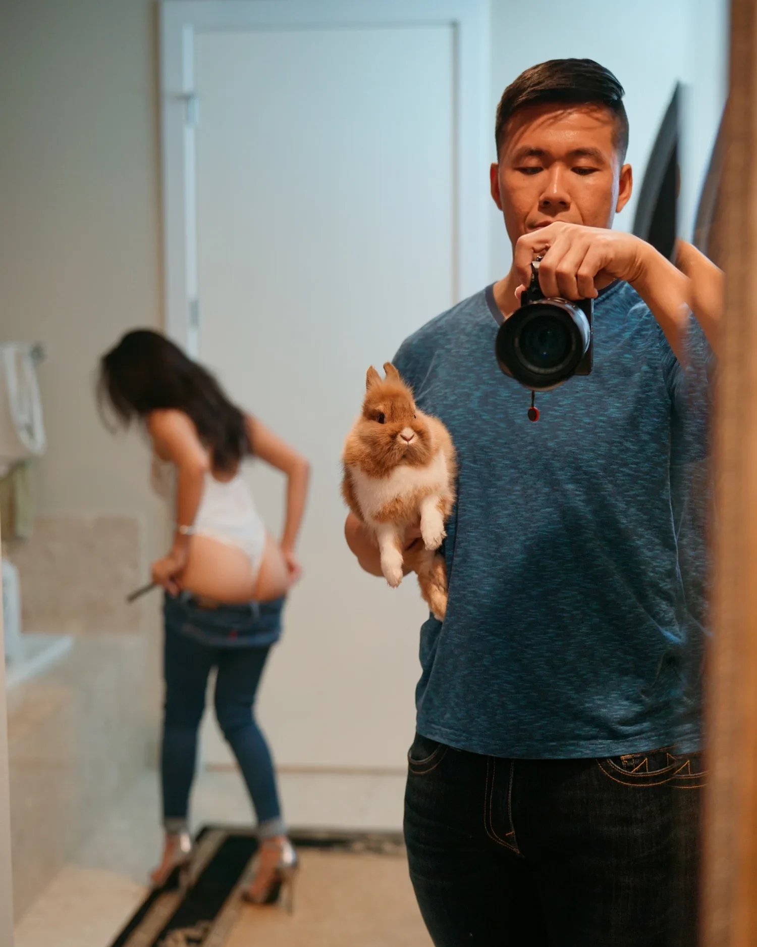 A man taking a mirror selfie with a camera, holding a fluffy bunny, while a woman in the background is adjusting her underwear.