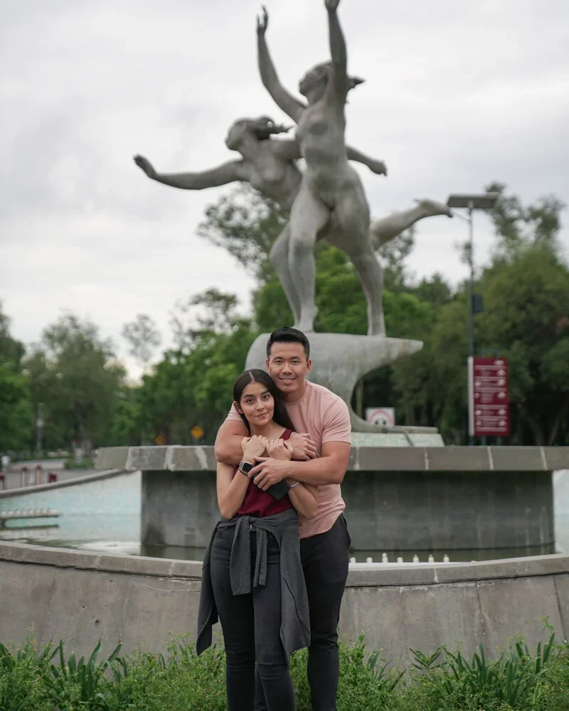 A young woman and young man hugging in front of a large outdoor sculpture of three intertwined figures with outstretched arms, in a park setting with green trees and cloudy sky.