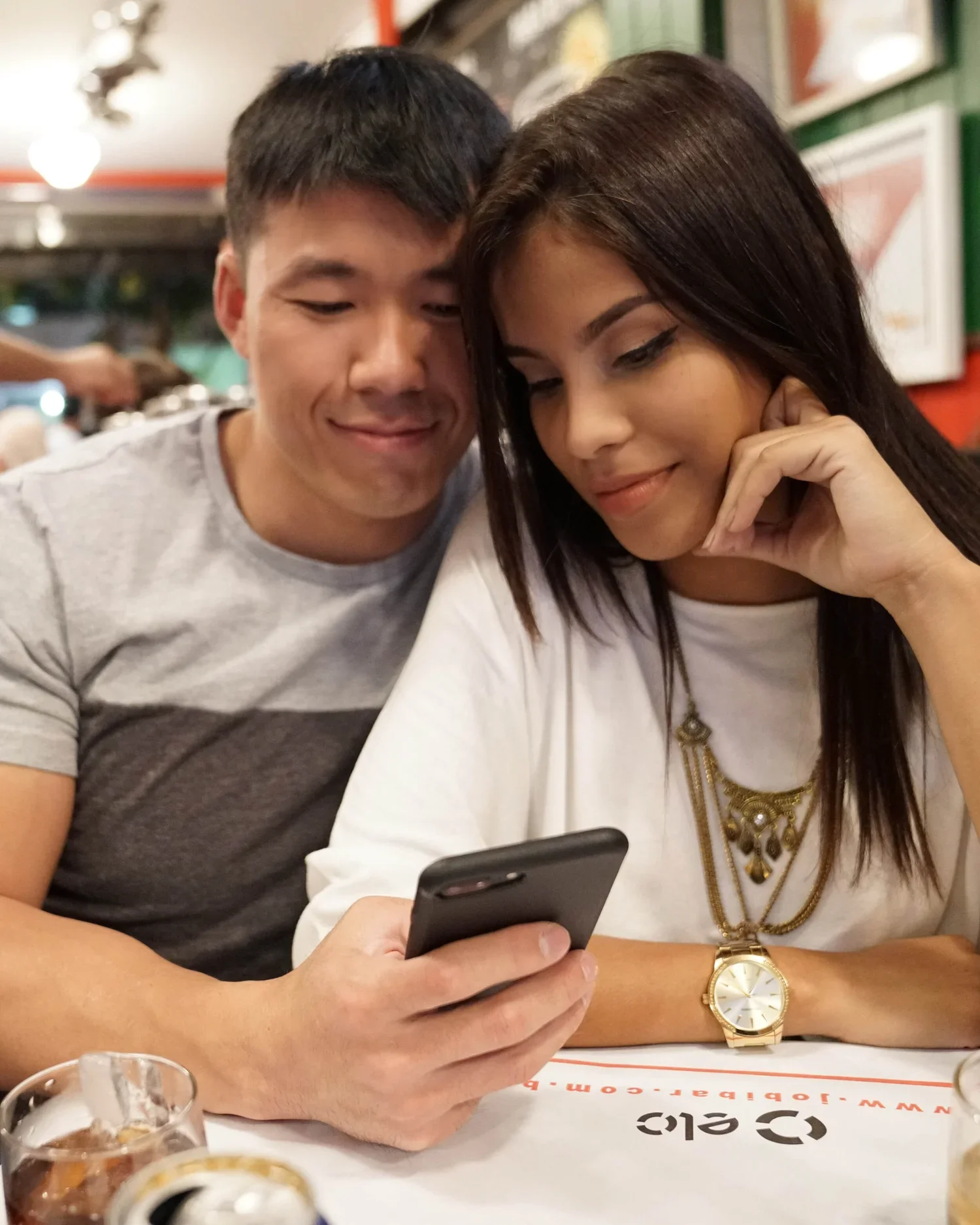 A young man and woman are sitting together at a table in a restaurant, looking at a smartphone and smiling.