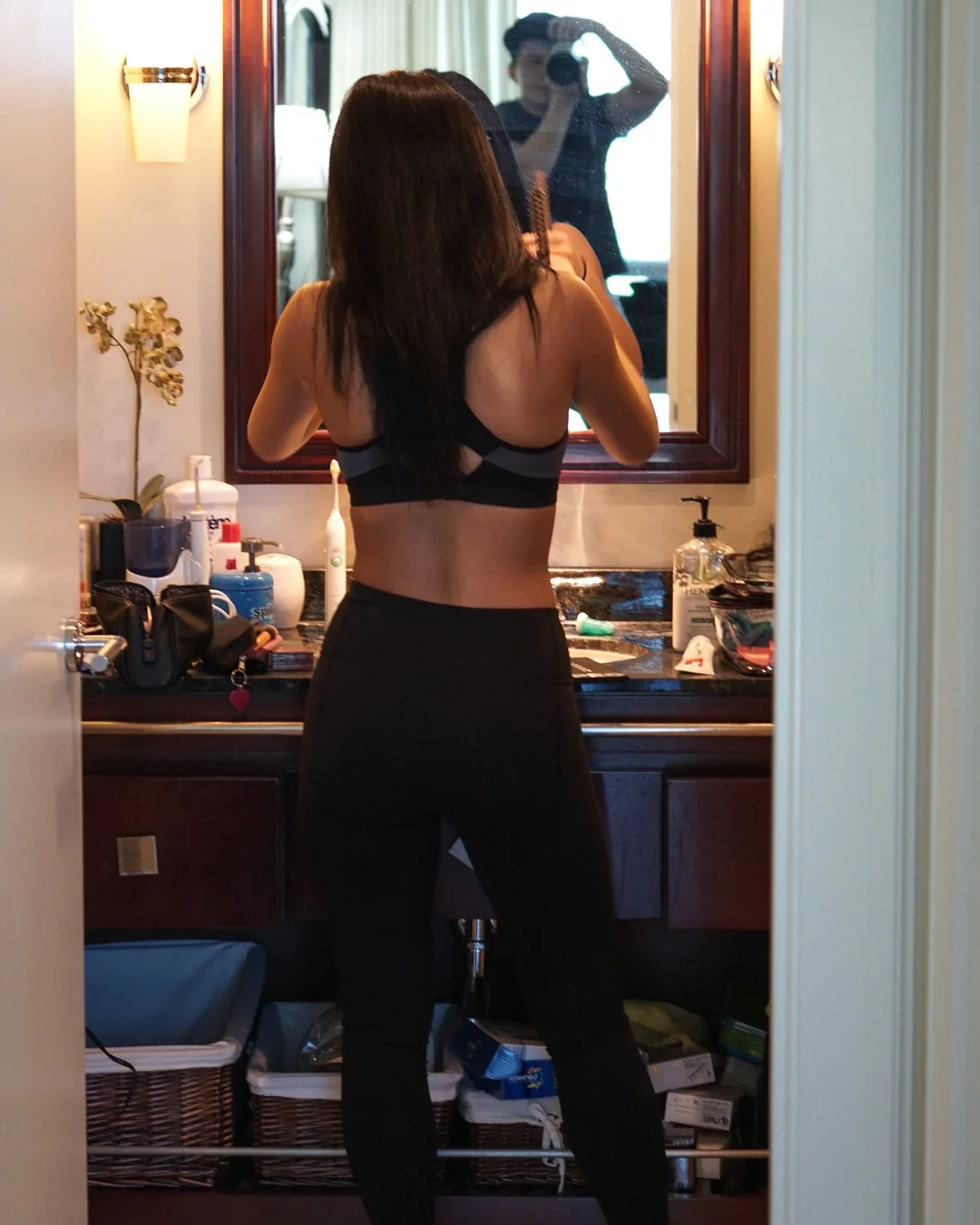 A woman with long dark hair takes a mirror selfie in a bathroom. She is wearing a black sports bra and black pants. The bathroom counter has various toiletries and a hand soap dispenser. A man is seen taking the photo through the mirror.