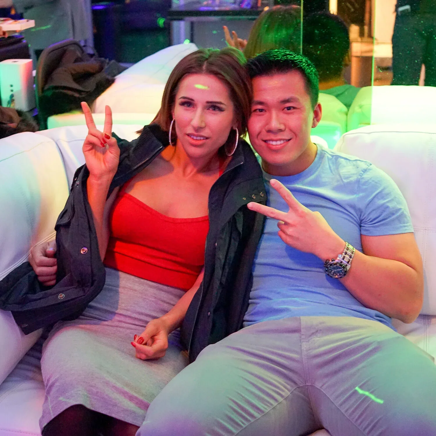 Two people sitting on a white couch at a nightclub, making peace signs and smiling at the camera. The woman has shoulder-length brown hair, hoop earrings, and is wearing a red top and black jacket. The man has short black hair, a light blue t-shirt, 