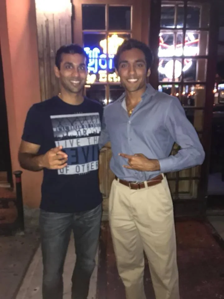 Two men standing side by side outside a restaurant or bar at night, smiling at the camera; one holding a drink, the other making a gesture with his hand.