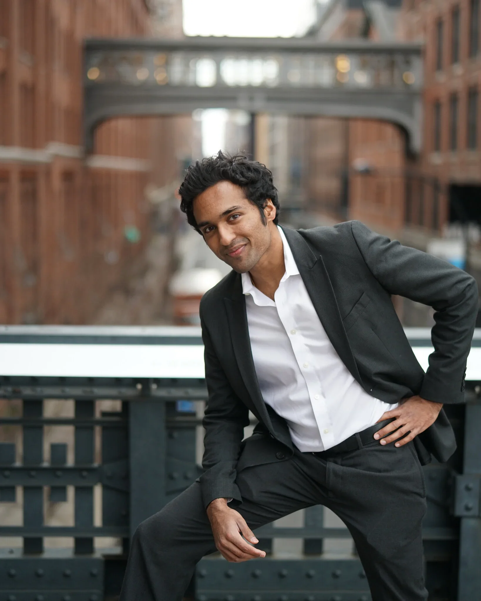 A man in a black suit with a white shirt, leaning forward with one hand on his knee and the other on his hip, smiling outdoors in an urban setting with brick buildings and a bridge in the background.