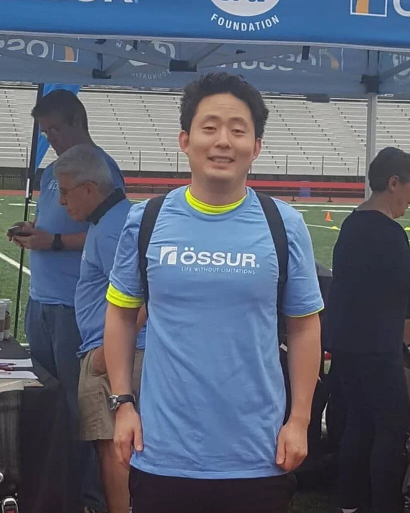 A young man with short black hair smiling, wearing a light blue sports shirt with the text 'Össur' and the slogan 'Life without limitations,' standing outdoors under a blue tent at a sports event, with a few other people in similar shirts and a stadium with empty bleachers in the background.
