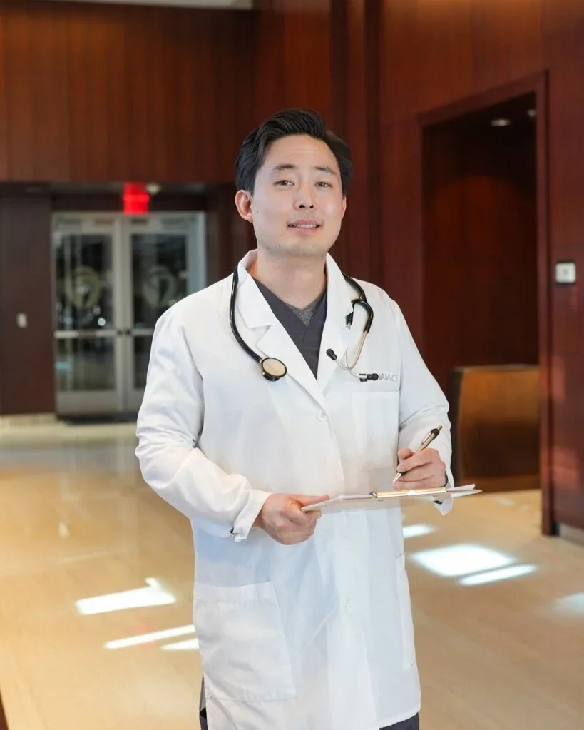 A male doctor in a white coat with a stethoscope standing in a hospital lobby, holding a clipboard and pen, with a confident expression.