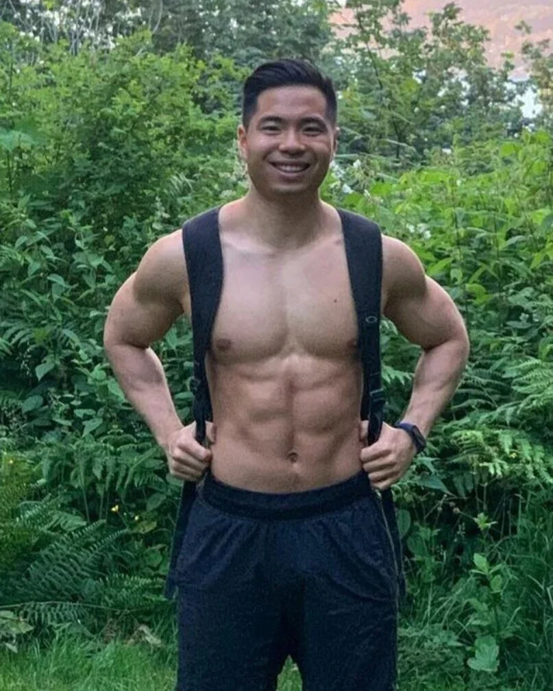 A shirtless man with short black hair, smiling, standing outdoors in front of green foliage, wearing black shorts and a backpack.