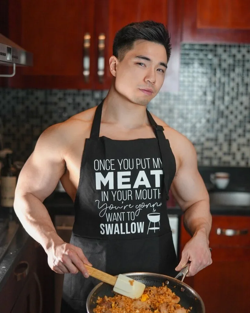 A shirtless man wearing a black apron with white text, standing in a kitchen and cooking fried rice in a pan.