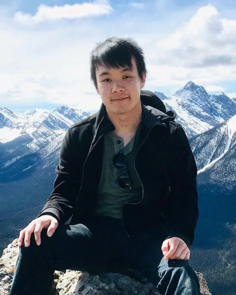 A young man sitting on a rock in front of snow-capped mountains under a partly cloudy sky.