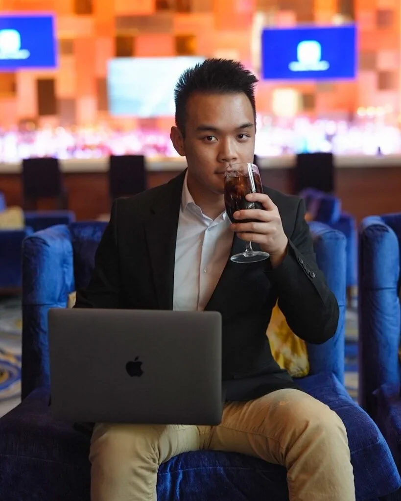 A young man in a black blazer and white shirt sitting on a blue couch in a lounge, holding a glass of soda and looking at the camera. There is an open MacBook on his lap and blurred screens in the background.