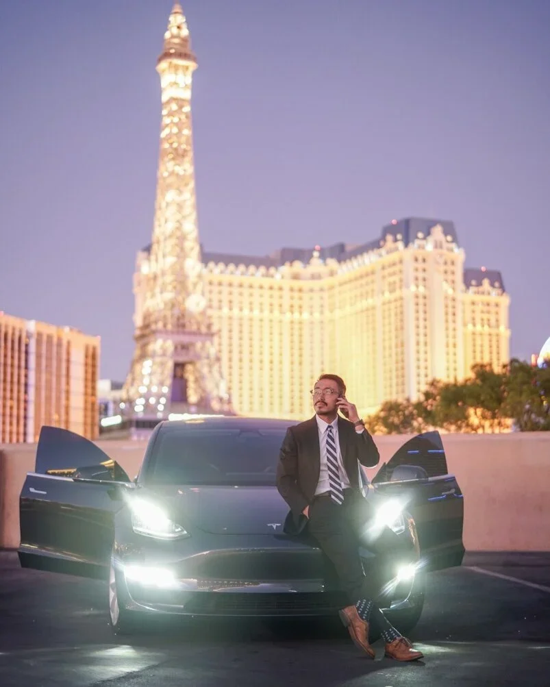 A man in a business suit leaning against a black luxury car with illuminated headlights, on a rooftop parking lot at night, with the Paris Las Vegas Hotel and the Eiffel Tower replica in the background.