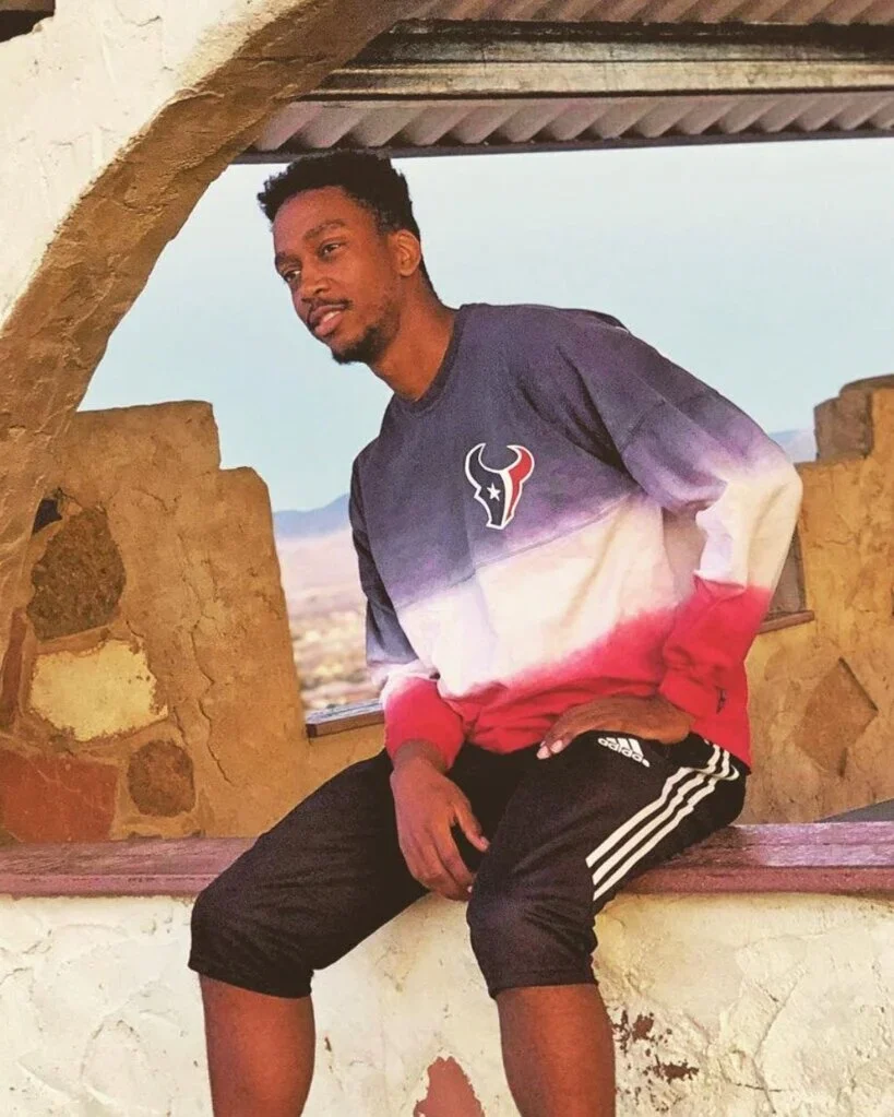 A young man sitting on a stone ledge near a window with a desert landscape in the background. He is wearing a Houston Texans sweatshirt and Adidas shorts.