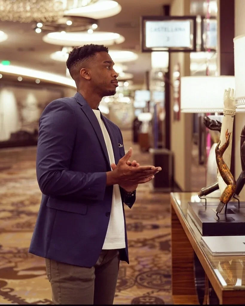 A young man in a blue blazer and white shirt looks at decorative sculptures in an upscale store or gallery, with warm lighting and a patterned carpet.
