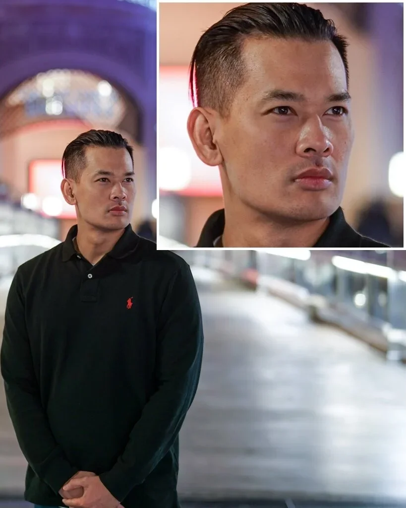 A man with short, slicked-back hair and a serious expression, wearing a black polo shirt with a red logo, standing in an indoor setting with blurred background and bright lighting.