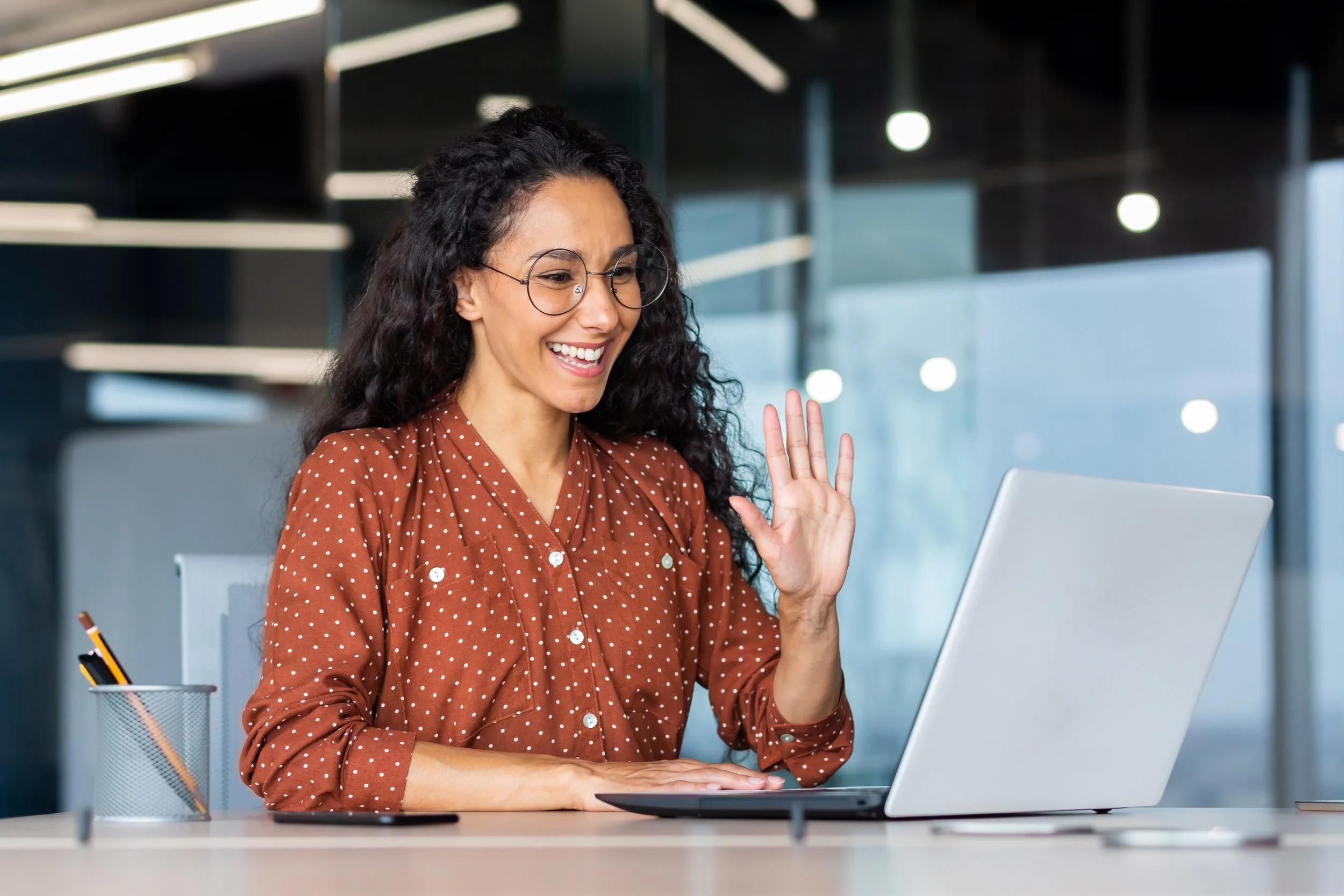 A woman with long curly hair and glasses smiling and waving at her laptop in a modern office.