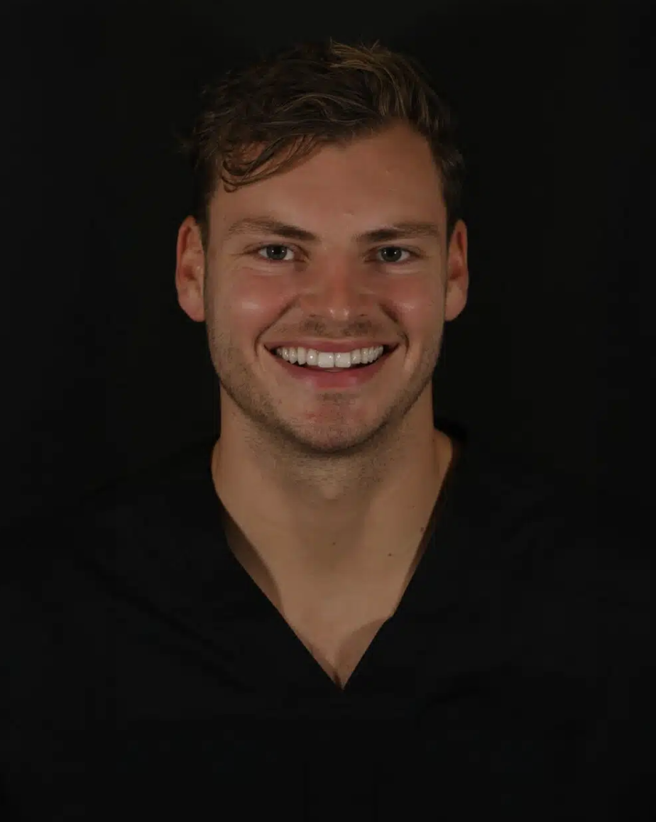 A young man smiling with short brown hair, wearing a black shirt, against a dark background.