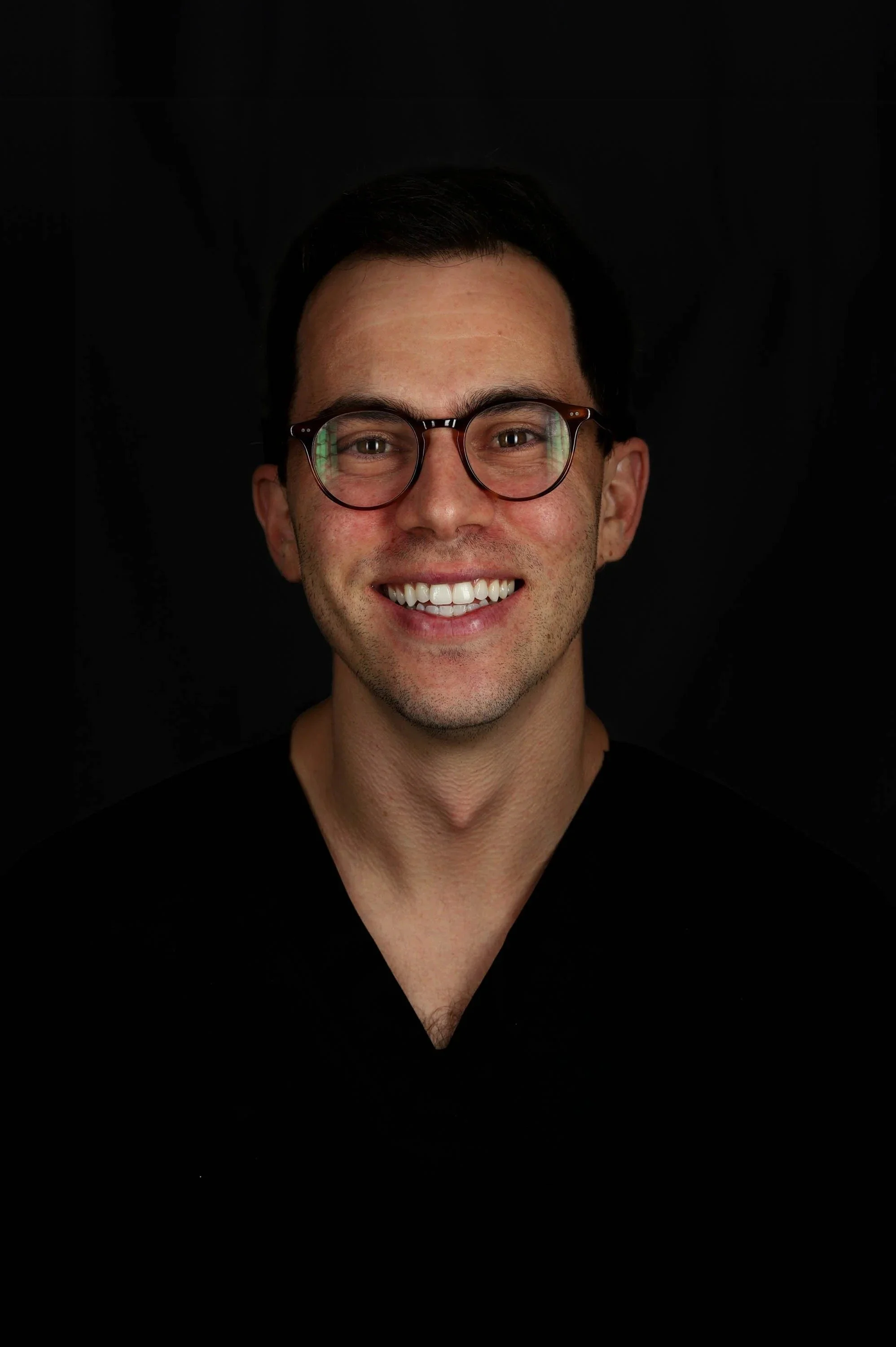 A smiling young man with dark hair, glasses, and a black shirt against a black background.