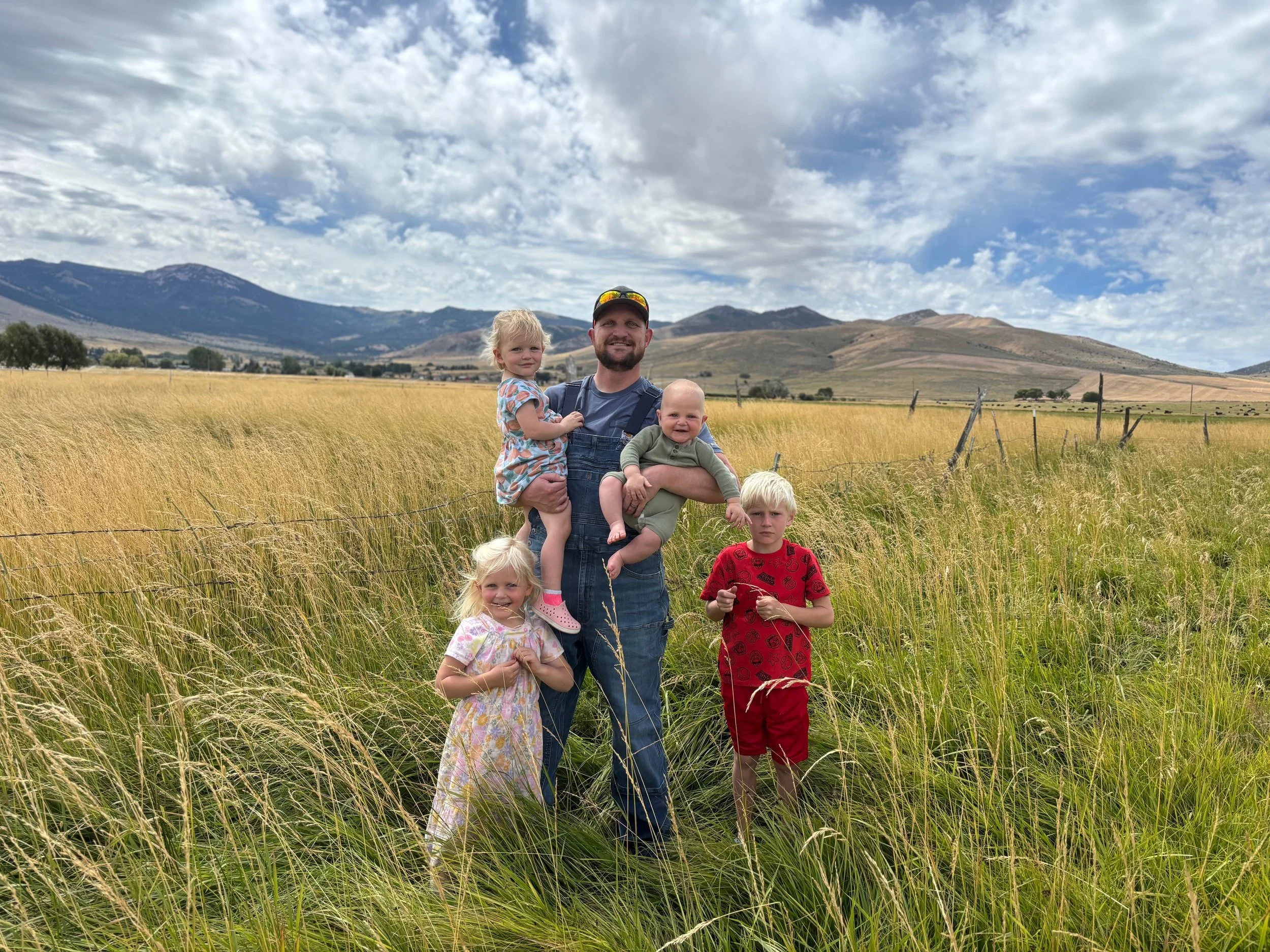 A man with four young children stands in a field of tall grass with mountains in the background under a cloudy sky.