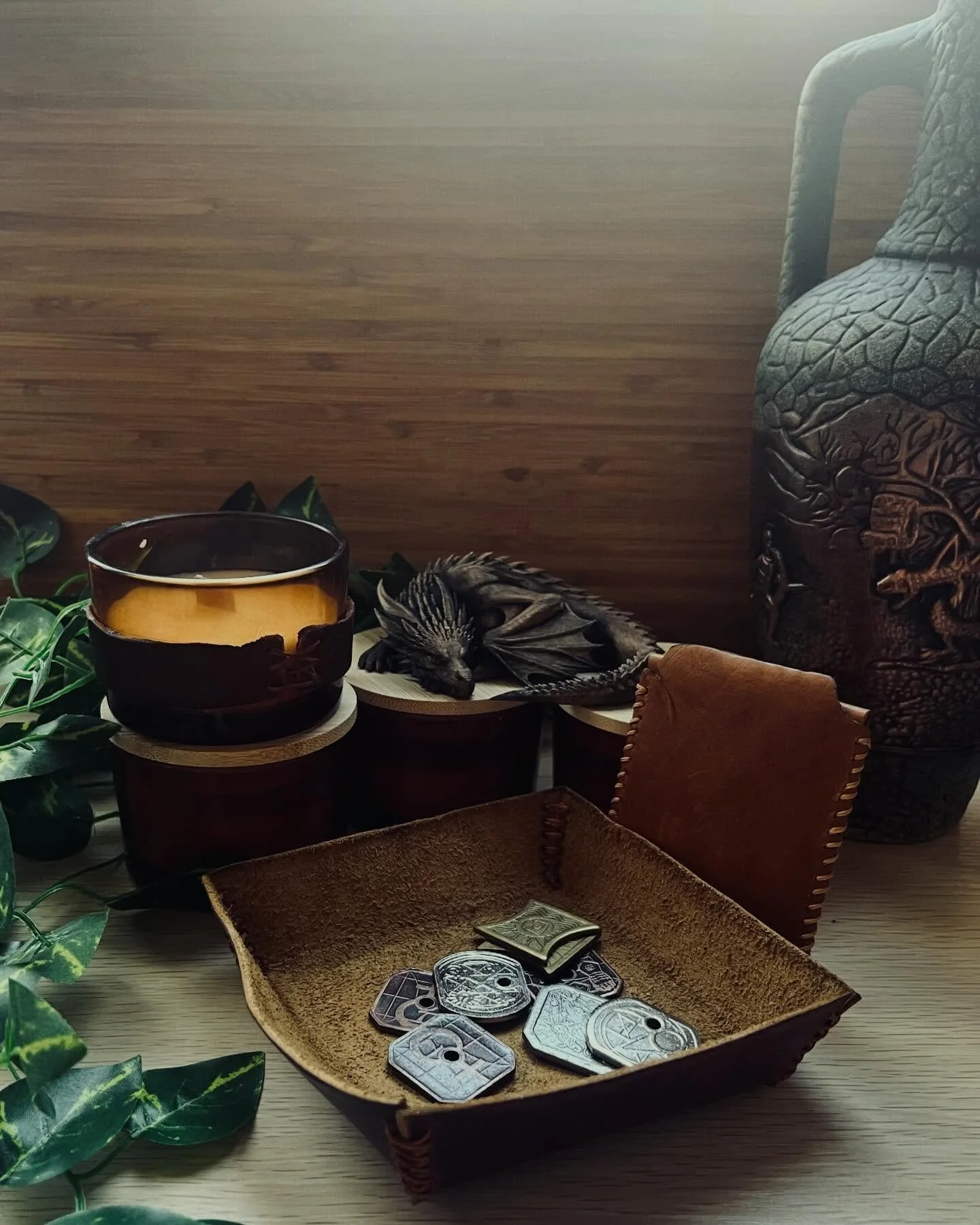 A cozy fantasy display with a hand-stitched leather dice tray, metal tabletop tokens, a warm candle, a sleeping dragon figure, greenery, and decorative dragon-themed décor.