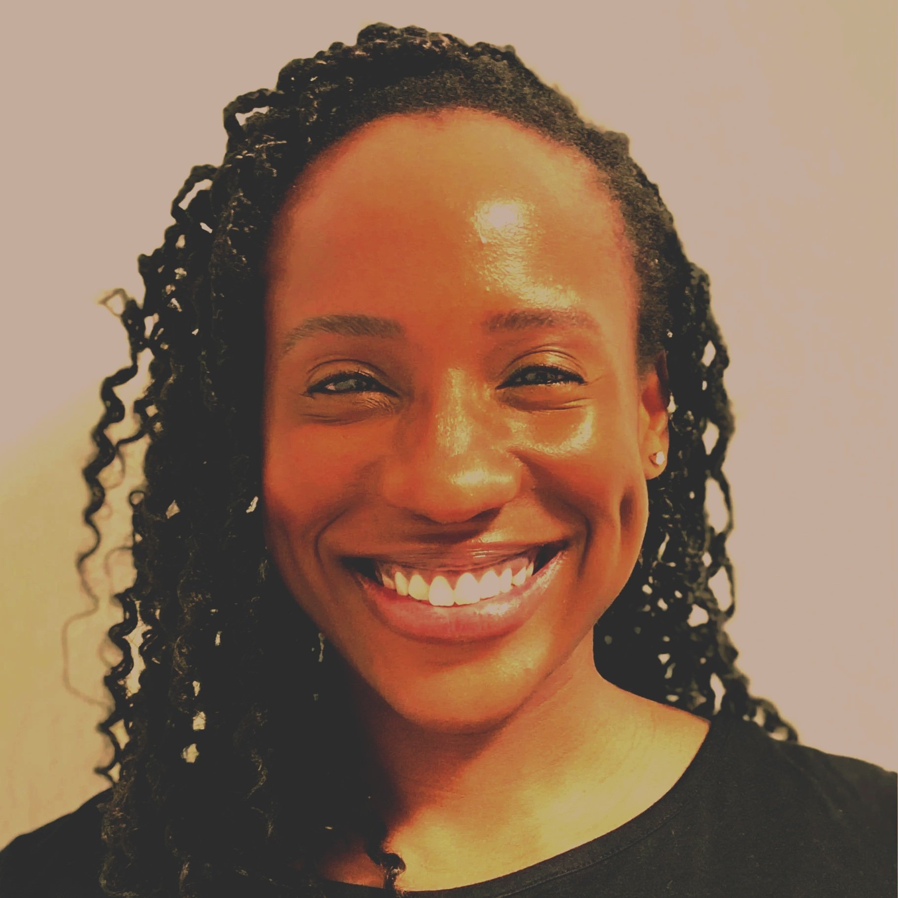 Close-up of a woman with dark curly hair smiling brightly, showing her teeth, against a plain background.