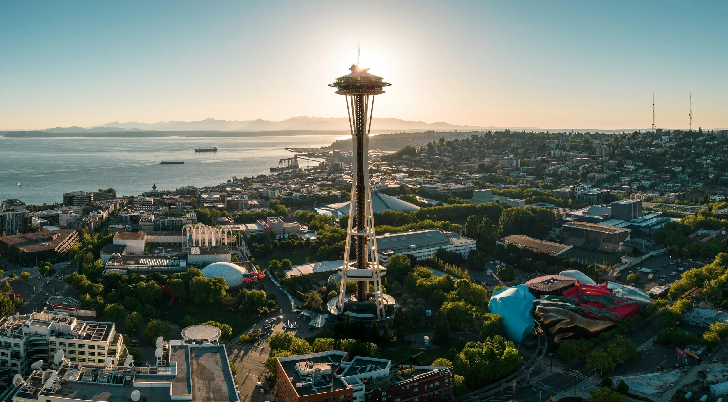 Aerial view of the Seattle skyline featuring the Space Needle in the foreground, with a waterfront, buildings, and mountains in the background during sunset.