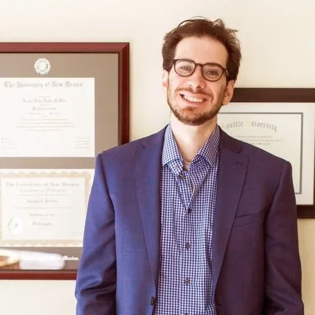Man with glasses and beard wearing a blue blazer and plaid shirt, smiling in front of framed diplomas or certificates on a wall.