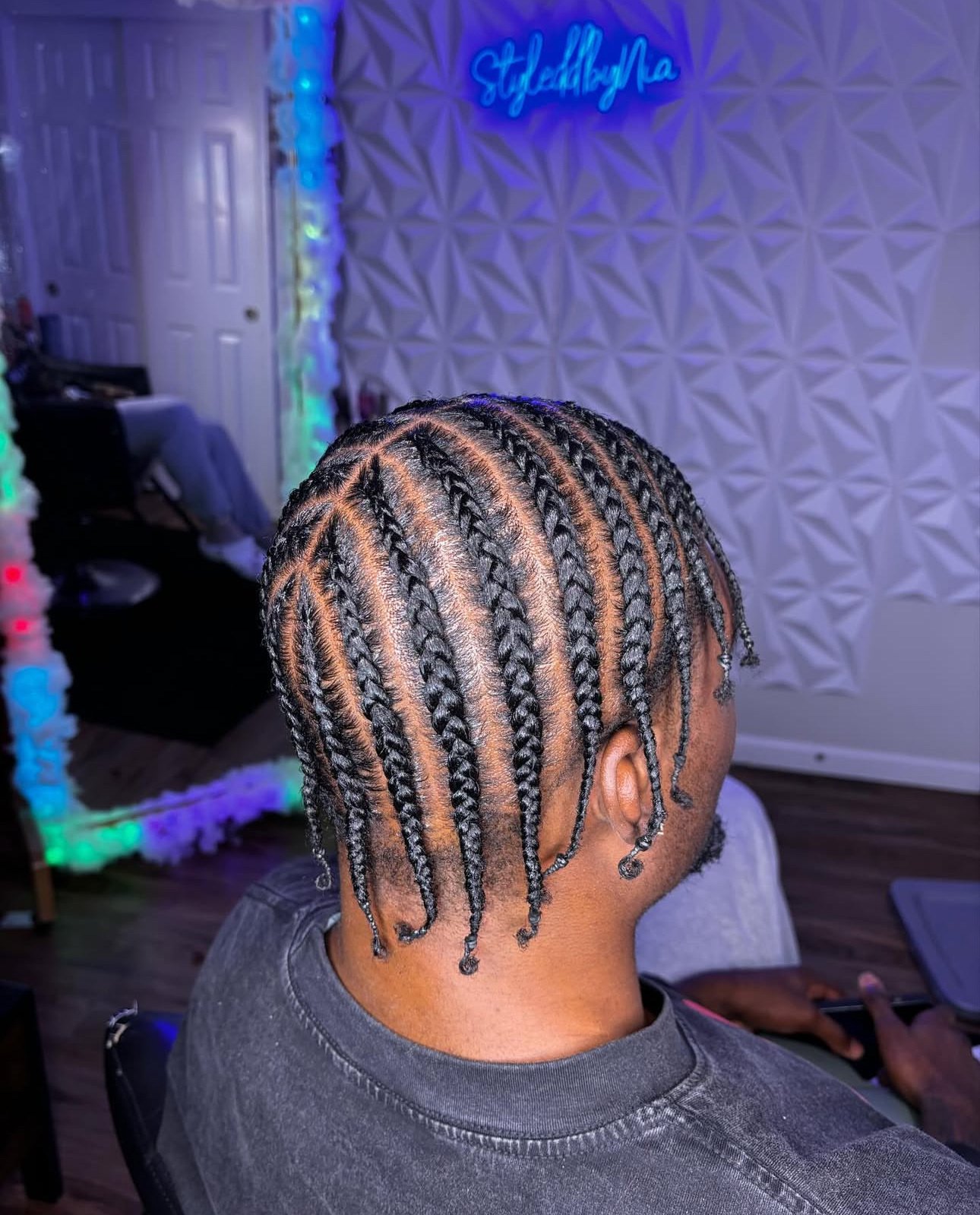A man with neatly styled cornrow braids in a room with decorative lighting and a neon sign on the wall.