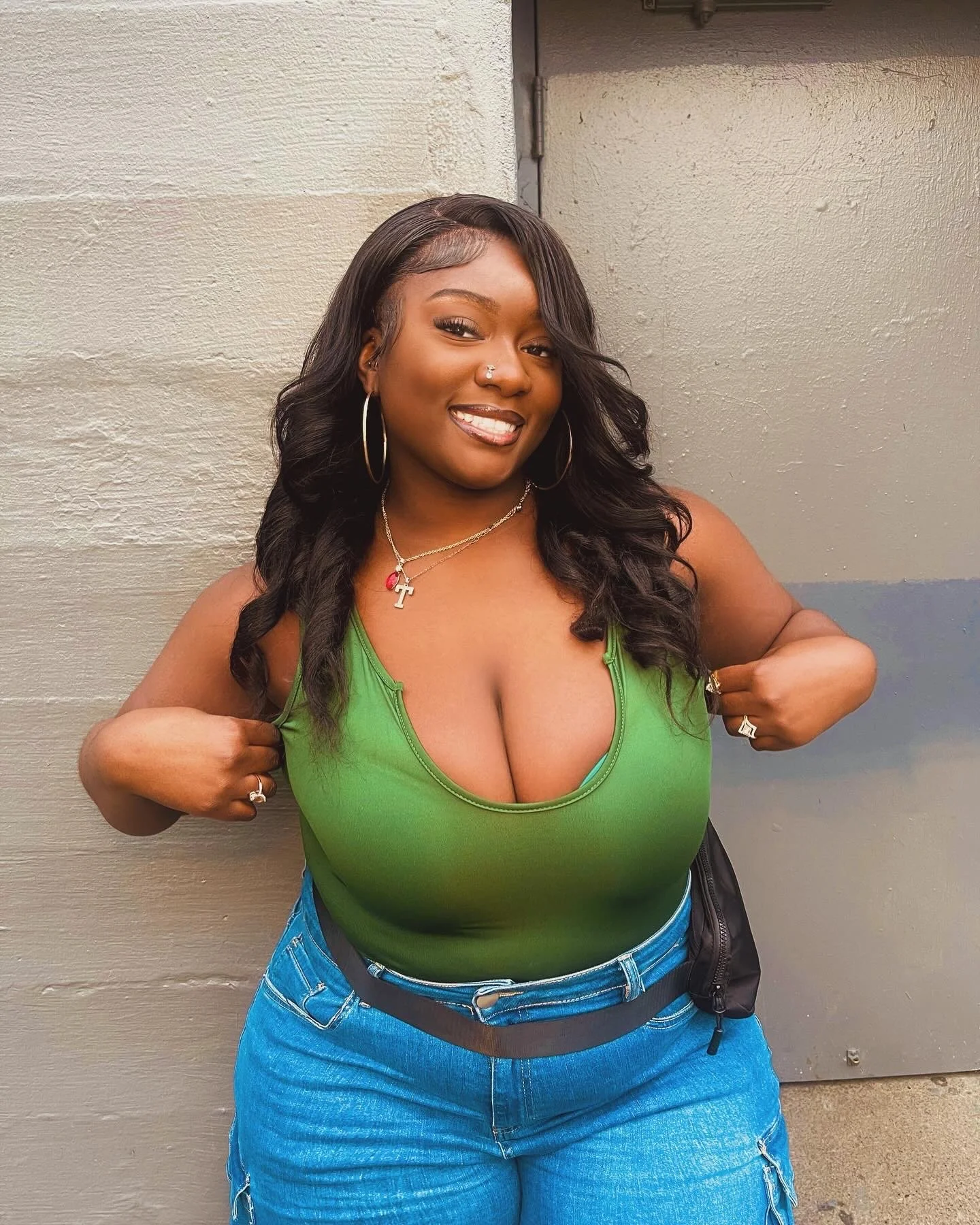 A smiling woman with long black wavy hair, hoop earrings, and a nose piercing, wearing a green tank top and blue jeans, standing against a gray wall.