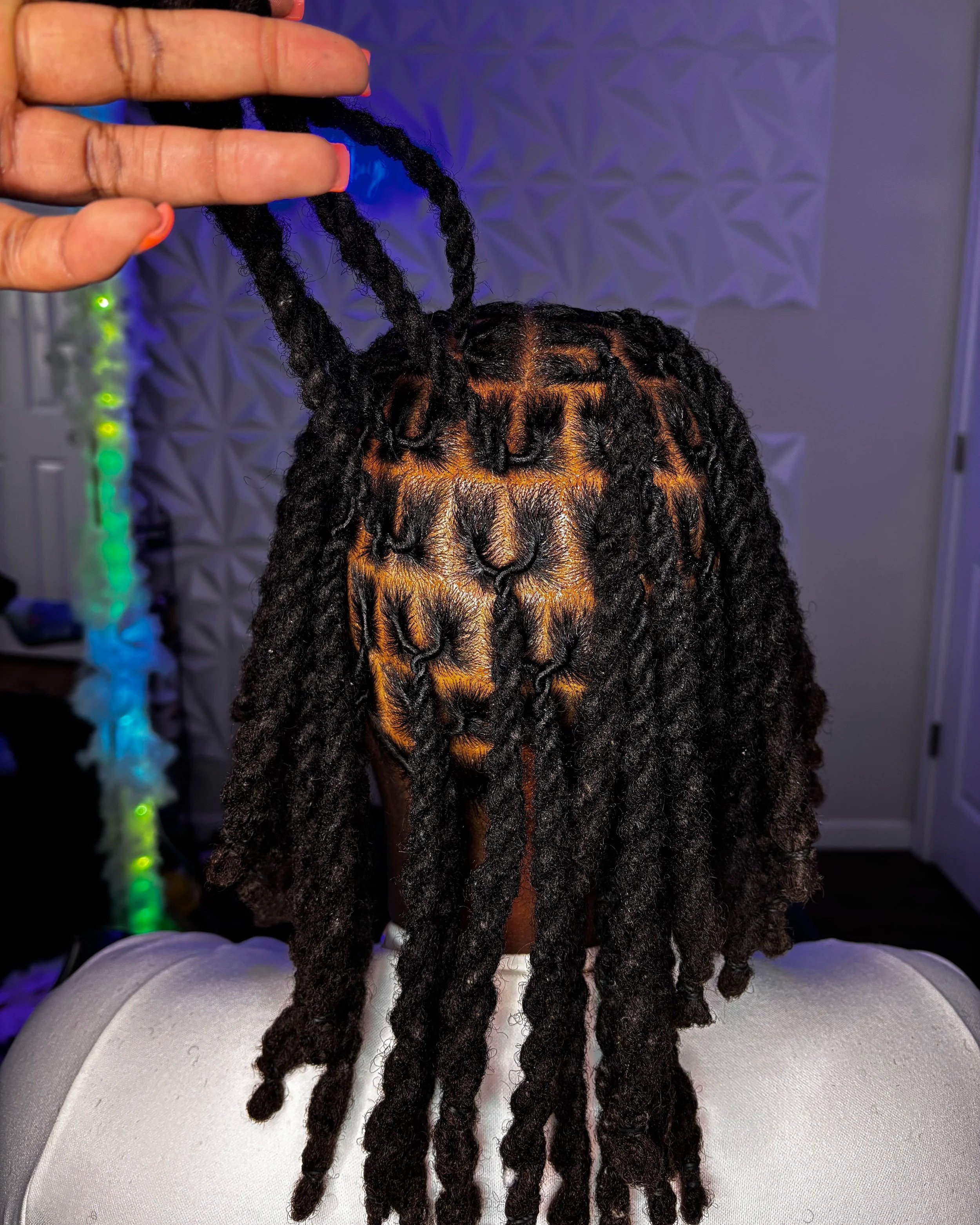 Person with styled dreadlocks, with one lock lifted by hand, in a room with gray textured wall and colorful LED lights.