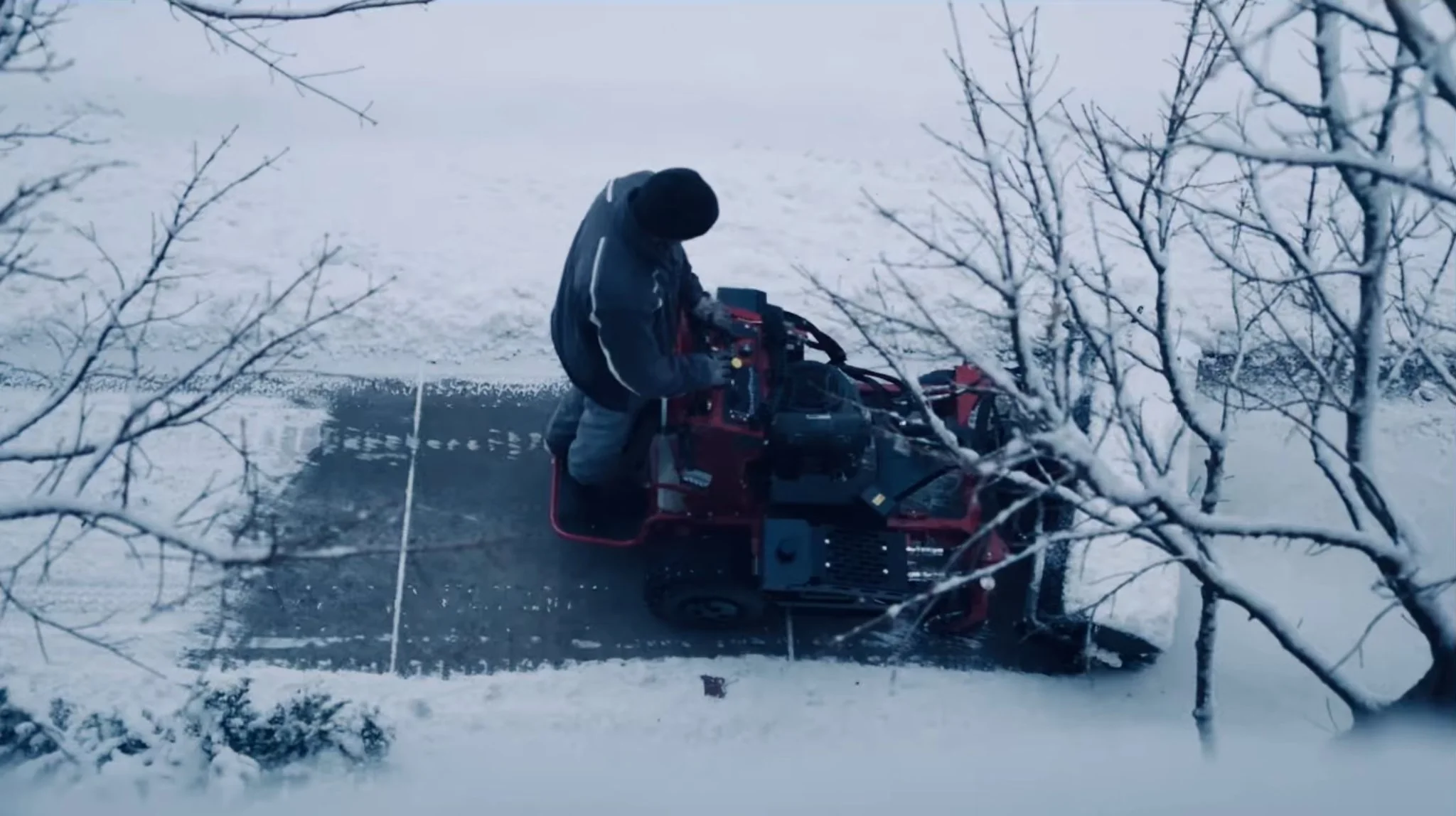 A person operating a snowblower on a snow-covered sidewalk, with leafless trees and snow in the background.