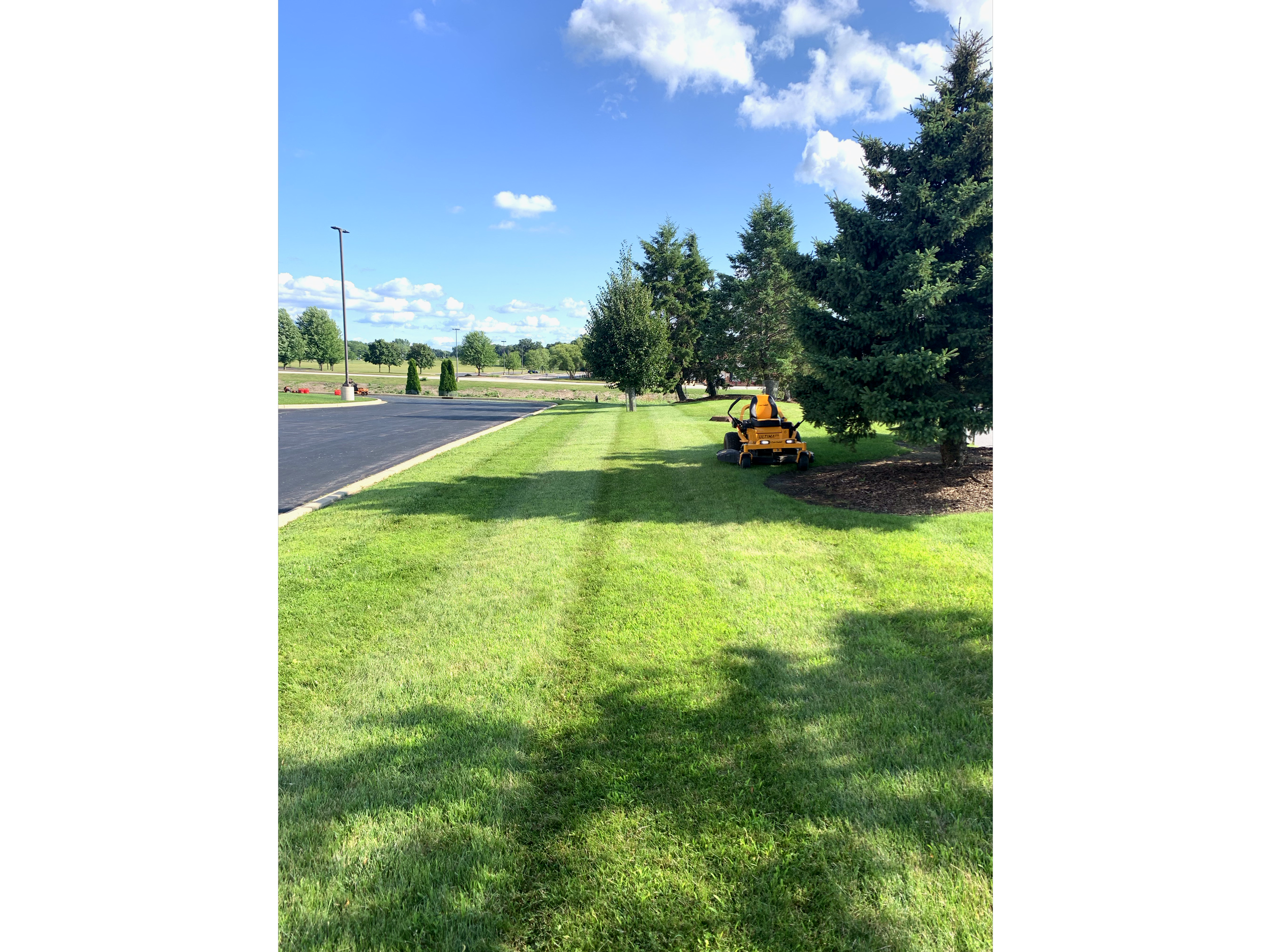 A yellow riding lawn mower parked on well-manicured green grass alongside large pine trees with a parking lot on the left and a blue sky with scattered clouds overhead.