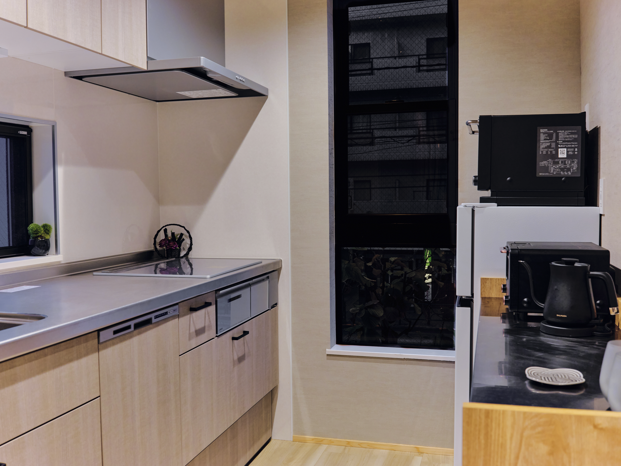 Modern kitchen with light wood cabinetry, a window with small potted plants, a stovetop with a vent hood, and a black microwave and kettle on the black countertop. A black-framed window shows a nighttime cityscape.