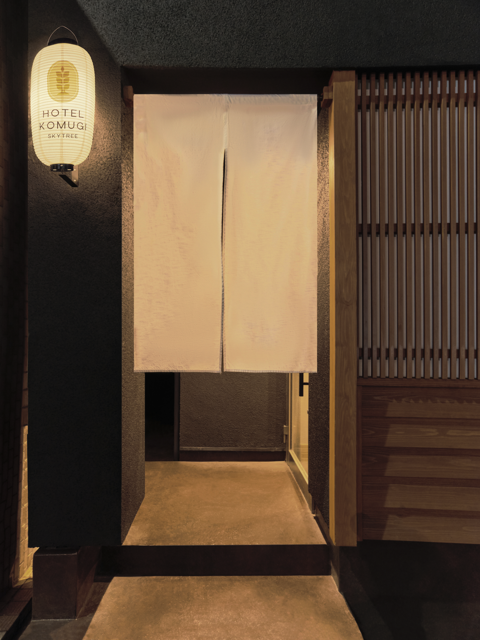 Entrance to a Japanese-style restaurant or hotel with a fabric noren curtain, wooden slatted wall, and a lantern that reads 'Hotel Komugi Skytree'.