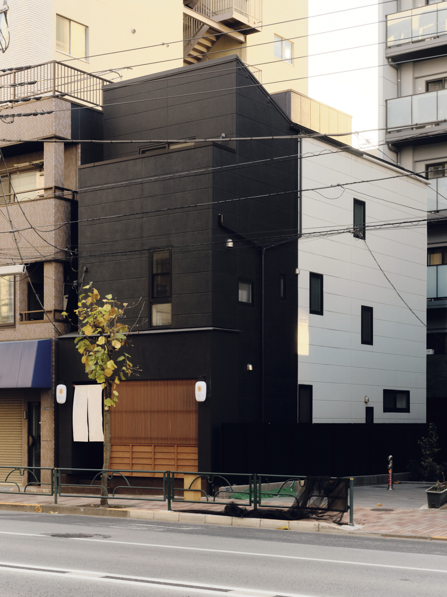 A modern building with black and white exterior walls, small windows, a wooden gate, and traditional Japanese lanterns outside, located along a city street.